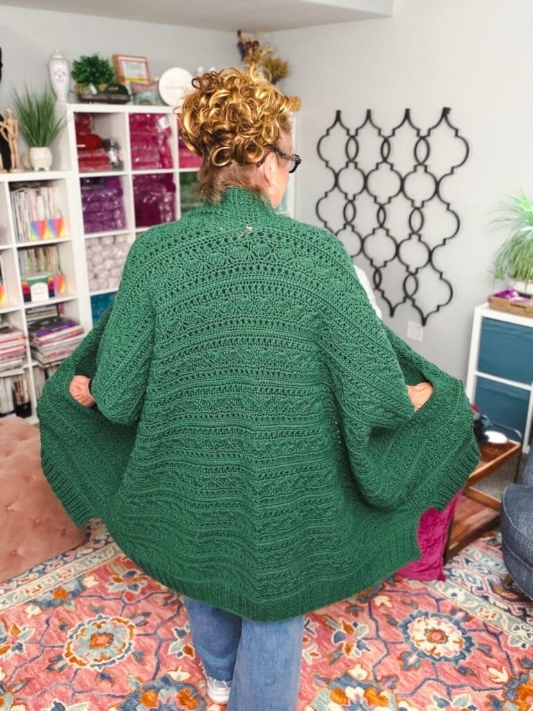 A textured green crochet ruana cardigan is modeled from the back, clearly showing stitch detail in a cozy, yarn-filled craft room.