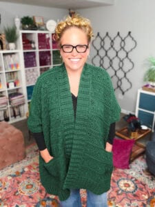 A person models a green crochet ruana cardigan with visible stitch detailing, shown worn over a black shirt in a cozy room.