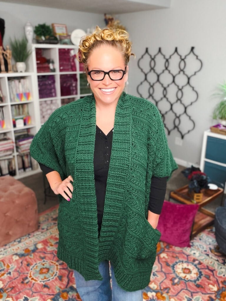 A green crochet ruana with visible stitch texture is modeled over a black top in a cozy room filled with yarn and books.