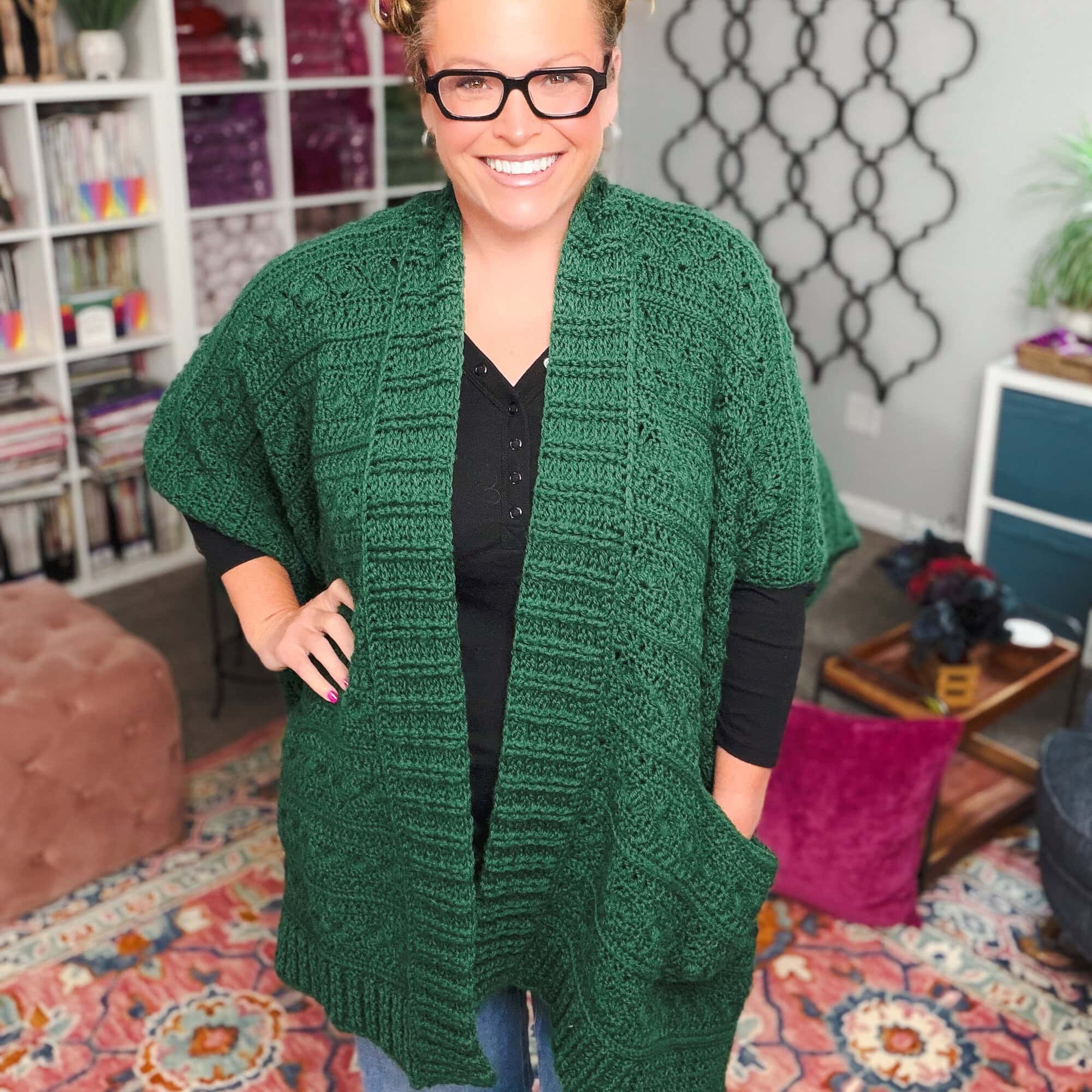 Person models a textured green crochet ruana-style cardigan over black top in a cozy, colorful room with patterned rugs.
