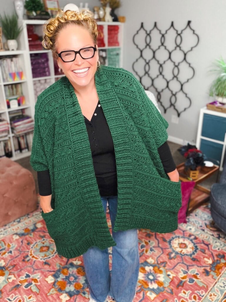 Green crochet ruana shawl draped over black top and jeans; stitch detail visible in a cozy, colorful room with bookshelves.