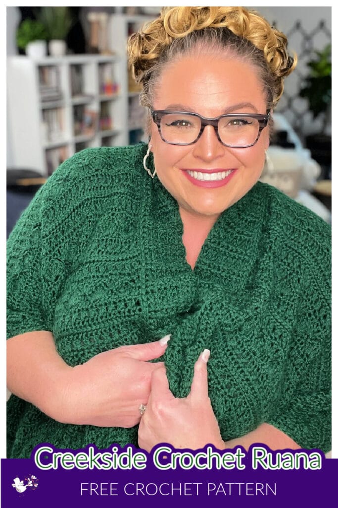 A smiling person wearing glasses and a green crocheted garment poses indoors, showcasing the cozy elegance of a crochet ruana pattern. Text at the bottom reads: "Creekside Crochet Ruana, Free Crochet Pattern." Books and plants add warmth to the background. -Marly Bird