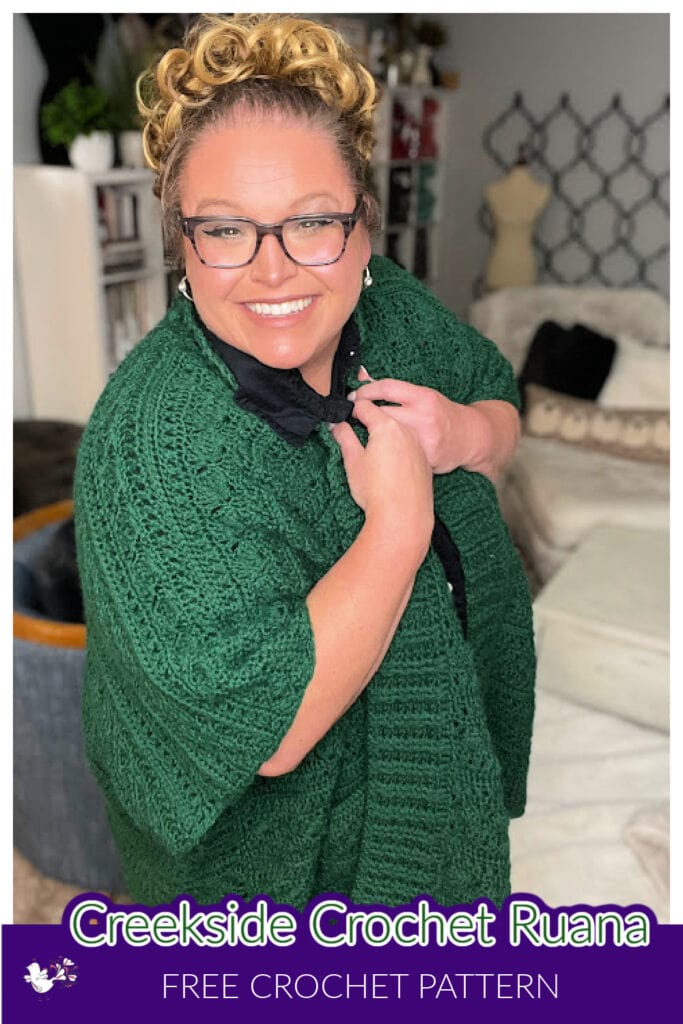 A person smiles while wearing glasses and a textured green crochet ruana, showcasing the latest crochet ruana pattern. The text at the bottom says "Creekside Crochet Ruana: Free Crochet Pattern." They stand indoors with casual decor in the background. -Marly Bird