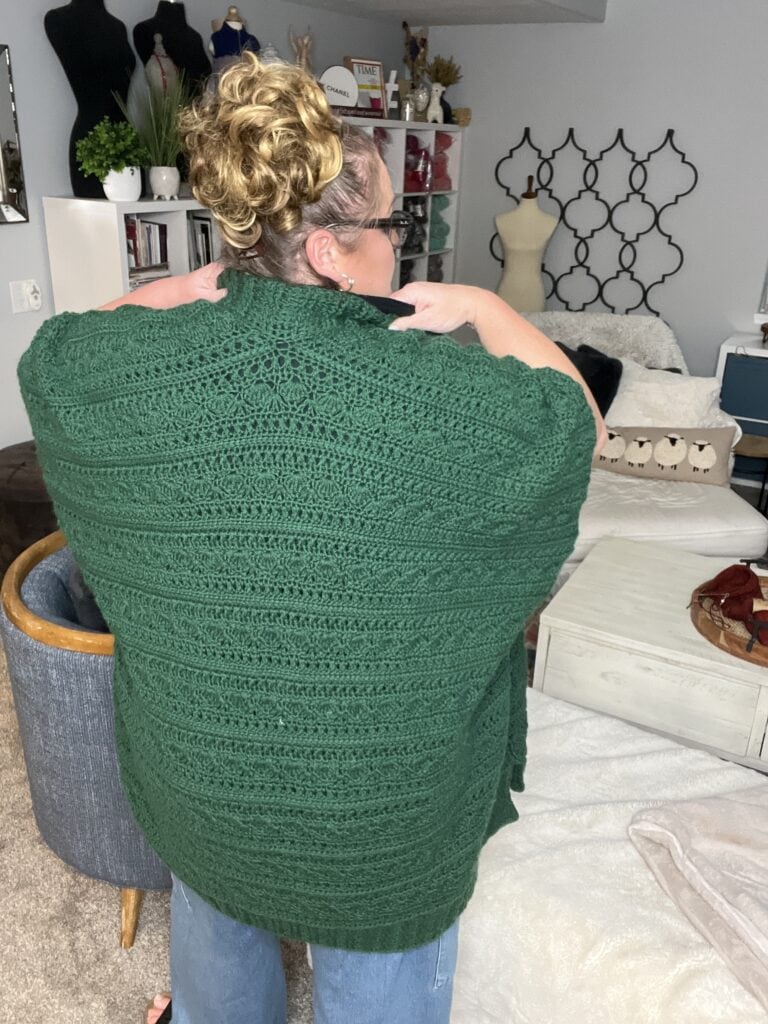 A person with curly hair stands with their back to the camera, showcasing a large green crocheted shawl designed in a beautiful ruana pattern. The room is cozy, adorned with bookshelves, a mirror, and various decorations in the background. -Marly Bird