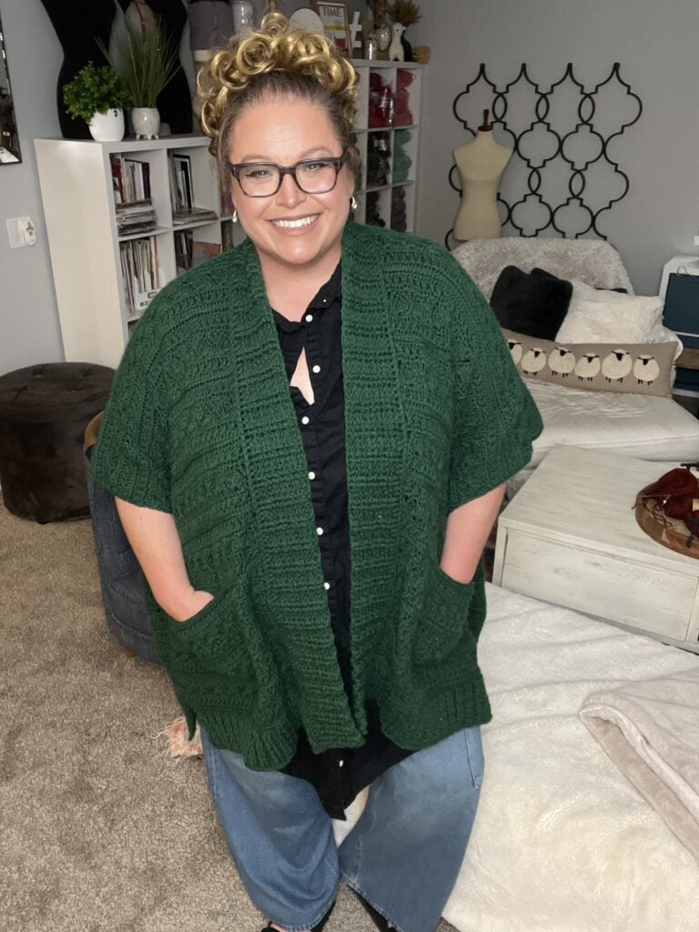 A person with curly blonde hair and glasses smiles while wearing a dark green knitted cardigan over a black shirt and jeans. The cozy space, adorned with home decor like a bookshelf and decorative wall piece, hints at inspiration for a crochet ruana pattern. -Marly Bird