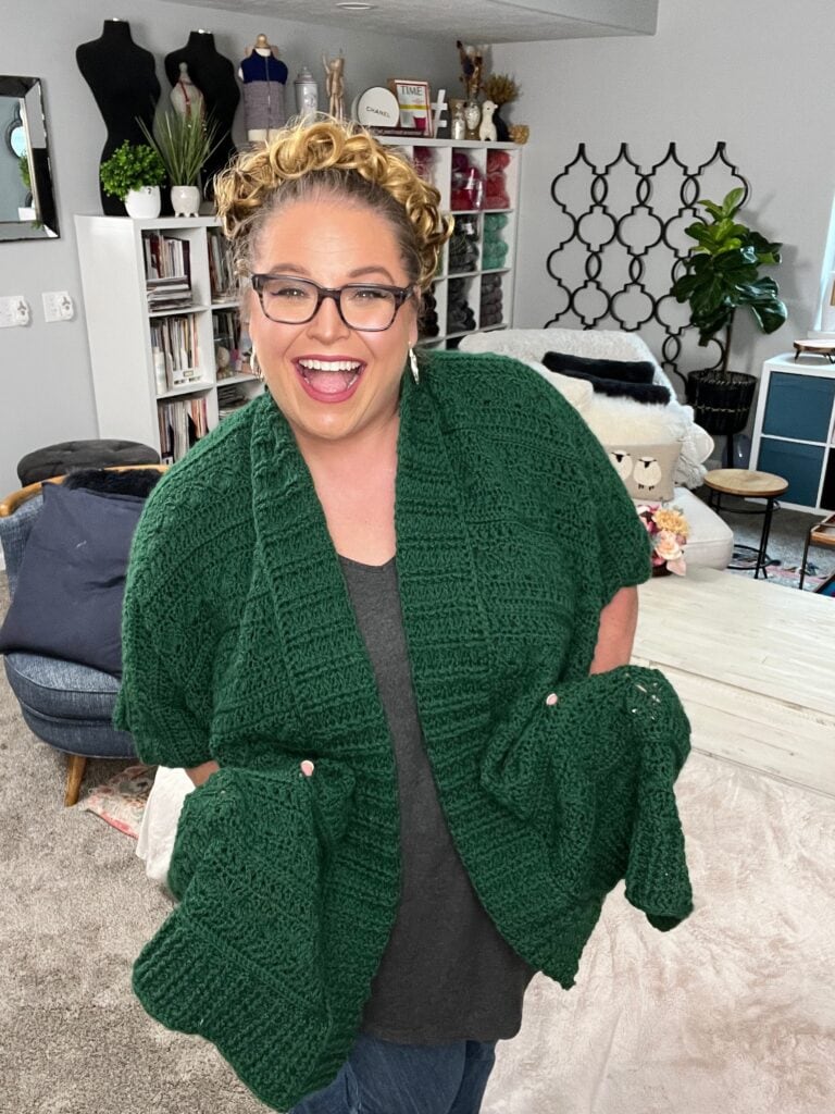 A person wearing glasses and a green crocheted shawl smiles broadly. They stand in a cozy room adorned with decor, bookshelves, and mannequins showcasing an array of crochet ruana patterns in the background. -Marly Bird