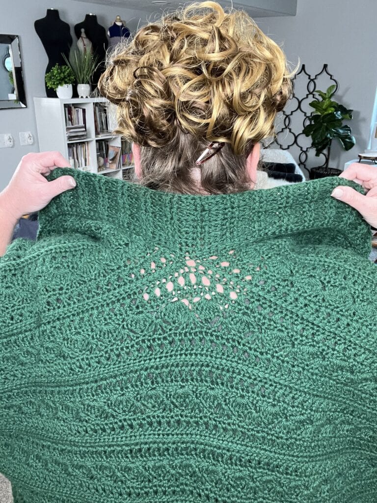 A person with curly hair seen from behind holds up a green knit shawl with intricate detailing that resembles a crochet ruana pattern. The background features bookshelves, a plant, and dress forms. -Marly Bird