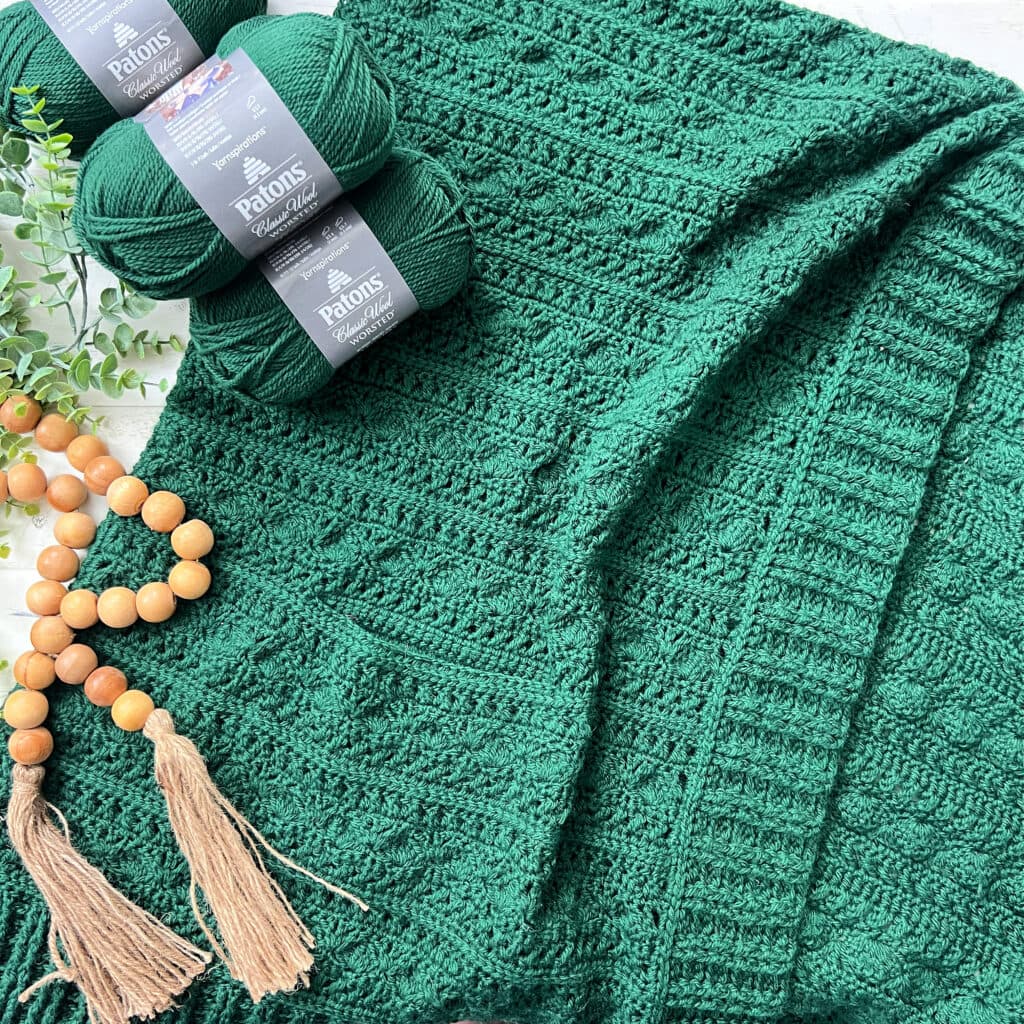 A textured green crocheted blanket is laid out with three skeins of matching Patons yarn, a wooden beaded garland with tassels, and a sprig of greenery beside it—perfect inspiration for your next crochet ruana pattern. -Marly Bird