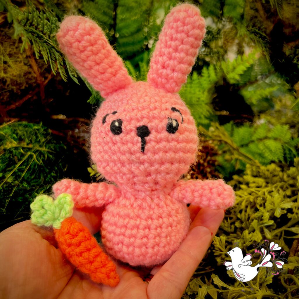 pink crochet bunny rabbit stuffed animal sitting on a hand in front of green background. Also has a crocheted carrot. Marly Bird logo in bottom right corner - pattern available on Marly Bird Website