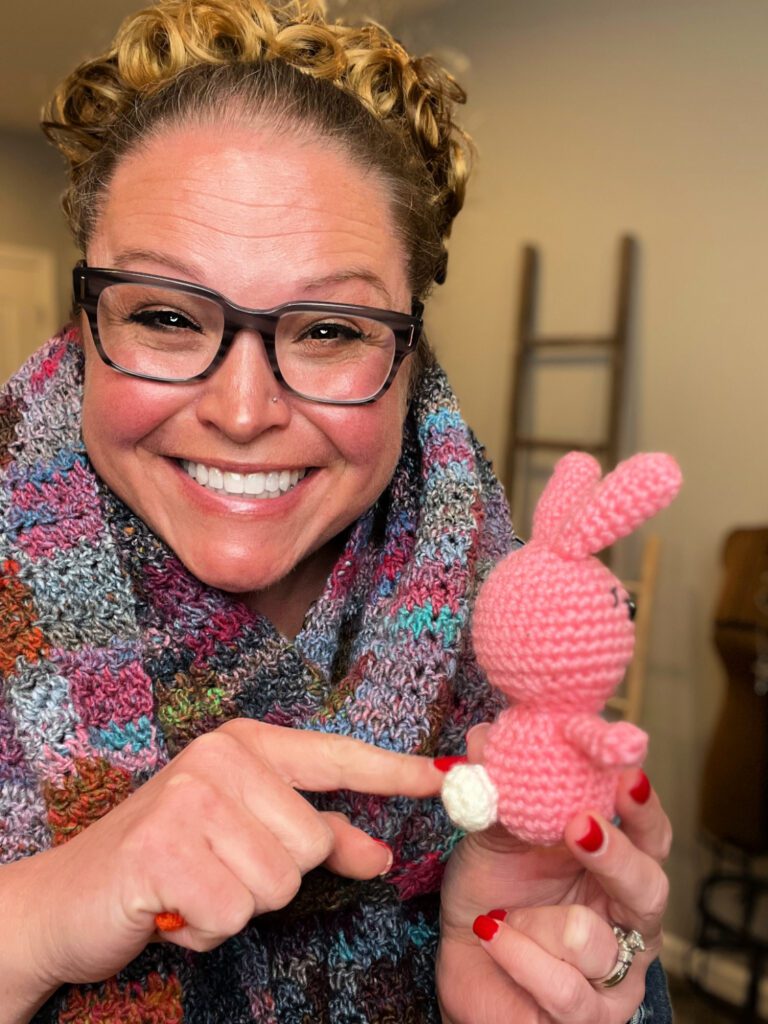 A beaming Marly Bird with curly hair and glasses showcasing a handmade pink rabbit. She is pointing at the white bunny tail.