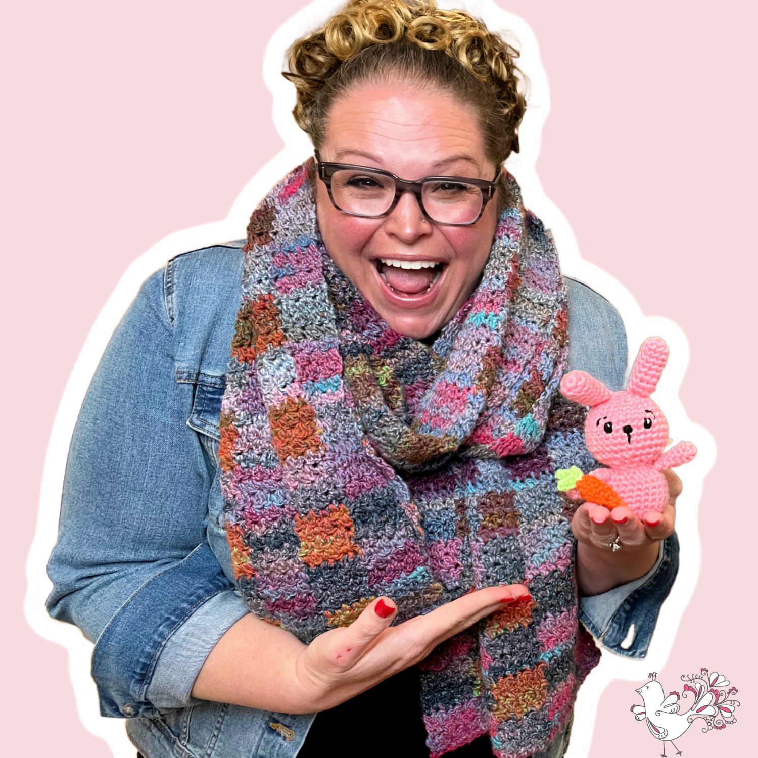 A joyous Marly Bird in a denim jacket and colorful scarf, beaming with delight as she presents a cute pink crocheted bunny rabbit with carrot. Free crochet bunny rabbit pattern on marlybird.com website