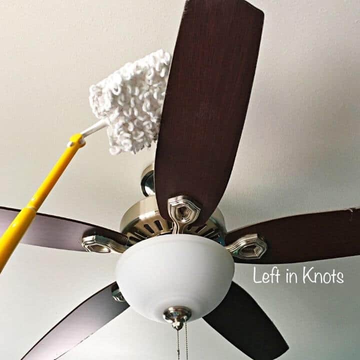 A brown ceiling fan with five blades is being dusted with a yellow-handled mop, perfect for spring cleaning. The fan features a central light fixture with a frosted glass cover. The mop's fluffy white head works efficiently against the plain white ceiling backdrop. -Marly Bird