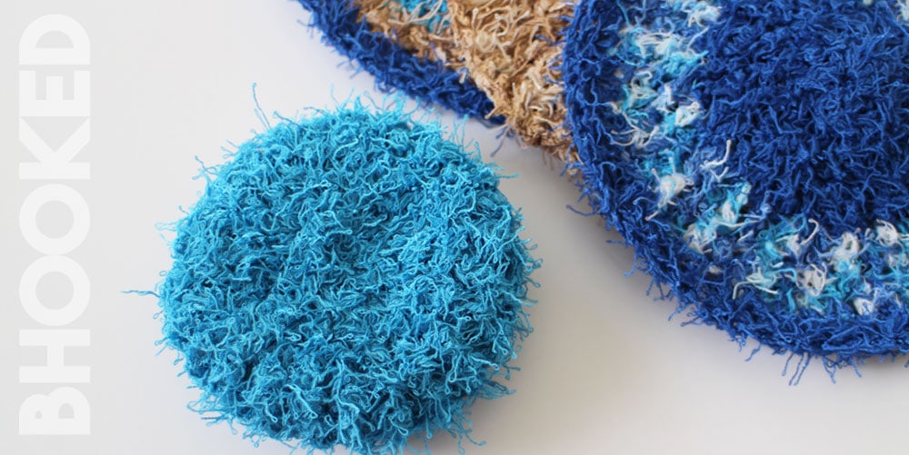 A close-up of crochet scrubbies: a round, textured blue scrubby in the foreground, perfect for your spring cleaning, with partially visible blue, white, and brown scrubbies in the background. The word "BHOOKED" is written vertically on the left. -Marly Bird