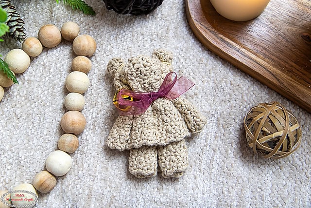 A crocheted teddy bear with a purple ribbon rests on a textured surface, embodying the freshness of spring. Nearby, wooden beads, a round wicker ball, and a wooden tray holding a candle complete the cozy and rustic scene. -Marly Bird