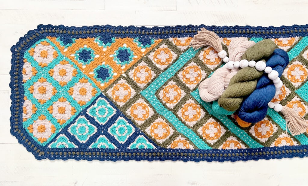 A colorful crocheted table runner with geometric and floral patterns in blue, teal, orange, and olive, bordered by dark blue. Several skeins of yarn and a tassel rest on the runner—perfect inspiration for new crochet patterns from Marly Bird. -Marly Bird
