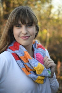 A woman with long brown hair and bangs is smiling at the camera. She is wearing a white top and a colorful, knitted scarf with red, orange, yellow, blue, and green sections. The background is a blurred outdoor setting with trees and sunlight. -Marly Bird