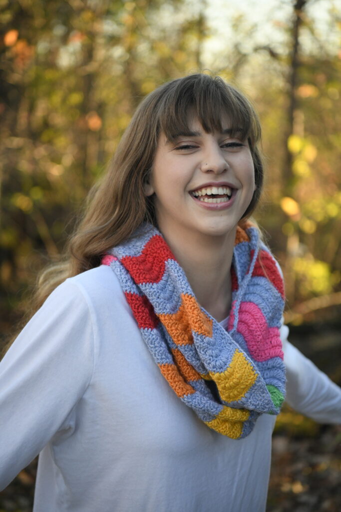 A person with shoulder-length brown hair is laughing and wearing a white shirt. They have a colorful, striped scarf around their neck. The background features blurred autumn foliage. -Marly Bird