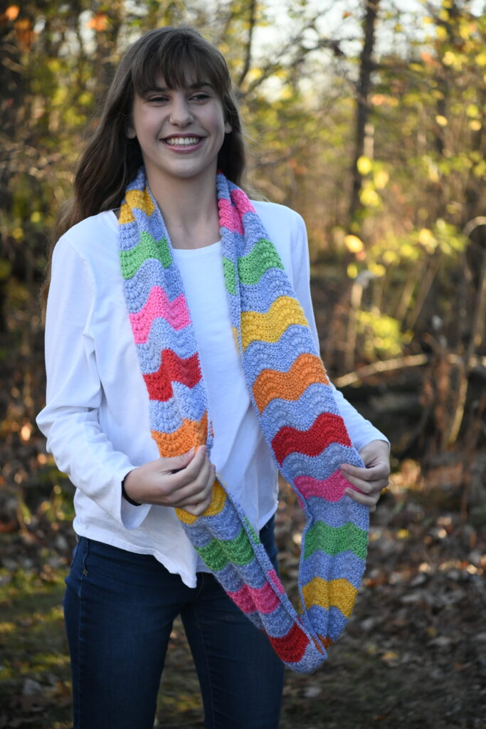 A person with long, brown hair is smiling and standing outdoors on a sunny day. They are wearing a white long-sleeve shirt and blue jeans. They are holding a colorful, striped crochet scarf in shades of red, orange, yellow, green, blue, and pink. The background features trees and foliage. -Marly Bird