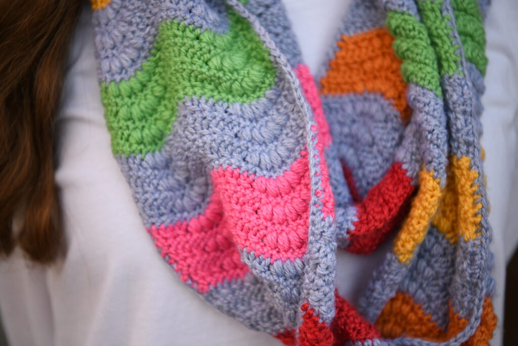 A close-up of a person wearing a crocheted scarf with a zigzag pattern in various colors including green, orange, pink, and grey. The scarf is draped loosely around their neck, and they are wearing a white garment underneath. -Marly Bird