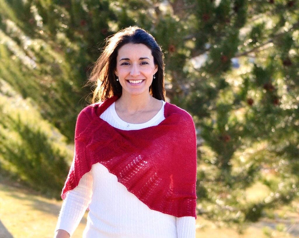 A woman models the Blood of My Blood shawl, showcasing its textured stitching and drape outdoors among green trees.