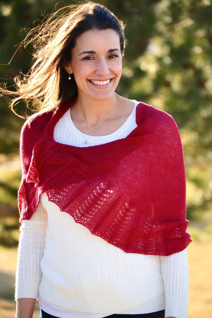 Bright red Outlander shawl in a textured knit drapes over a white sweater; worn by a woman outdoors with blurred greenery.