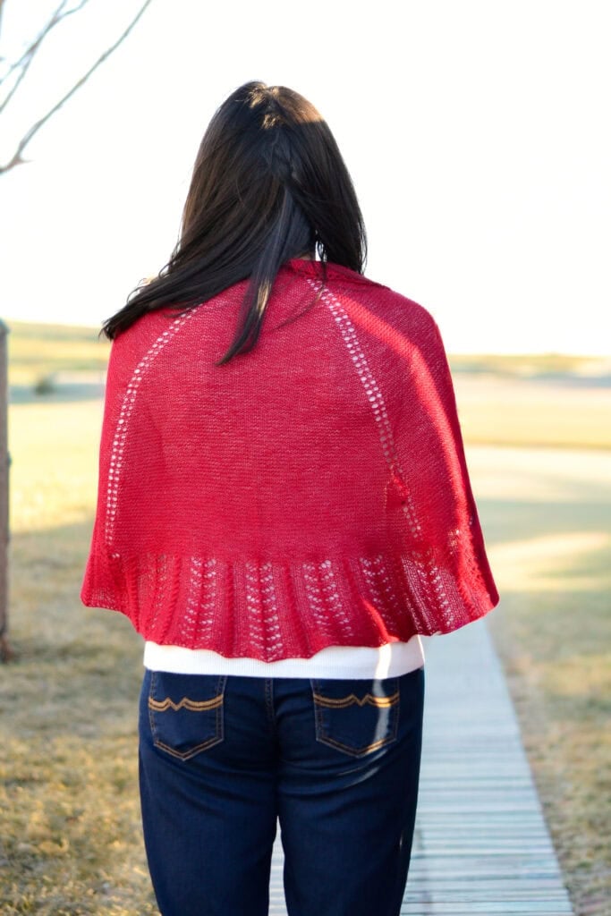 A bright red Blood of My Blood knit shawl with visible texture drapes over a person, displayed outdoors on a wooden path.