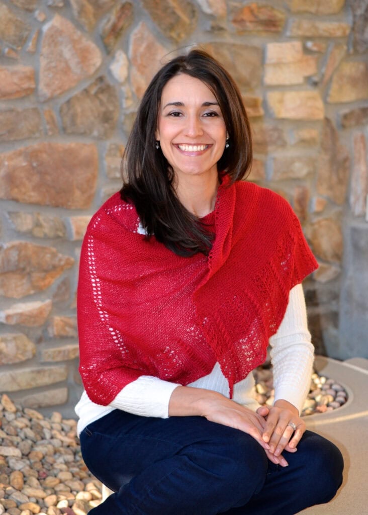 Woman models a red Blood of My Blood shawl, knit in textured yarn, over a white sweater, seated by a stone wall.