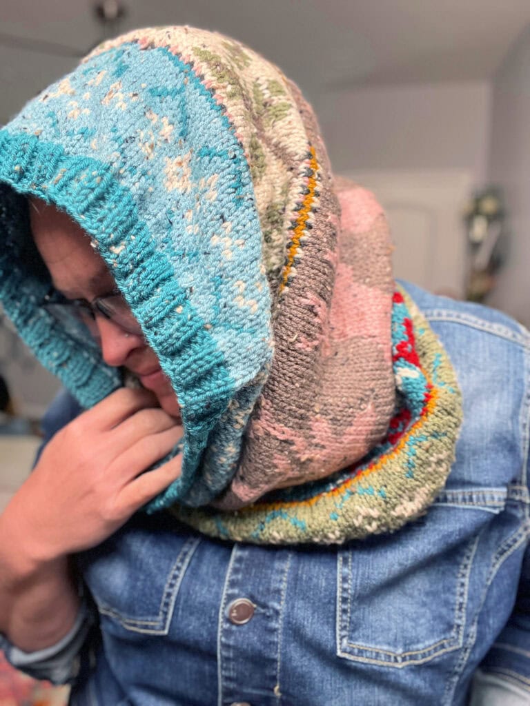 A person wearing glasses and a blue denim shirt is draped in a colorful, multi-patterned Dancer's Delight Knit Cowl, holding part of the knit cowl edging near their chin. The softly blurred background adds to the cozy allure. -Marly Bird