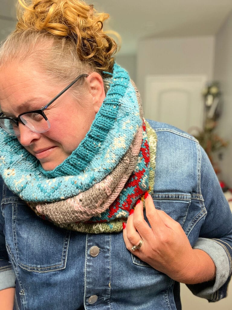 A person with light hair in a curly bun, wearing glasses, a denim jacket, and a colorful Dancer's Delight Knit Cowl looks down while holding the cowl. The background shows a softly lit indoor space with a door and some decor. -Marly Bird