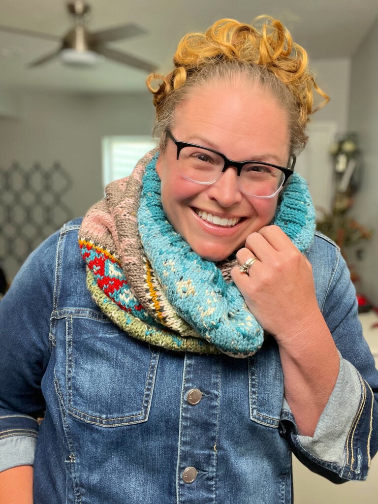 A smiling person with curly blonde hair, black glasses, and a denim jacket wears a vibrant Dancer's Delight Knit Cowl with detailed final edging, showing a ring on their left hand. The background features cozy indoor home decor. -Marly Bird