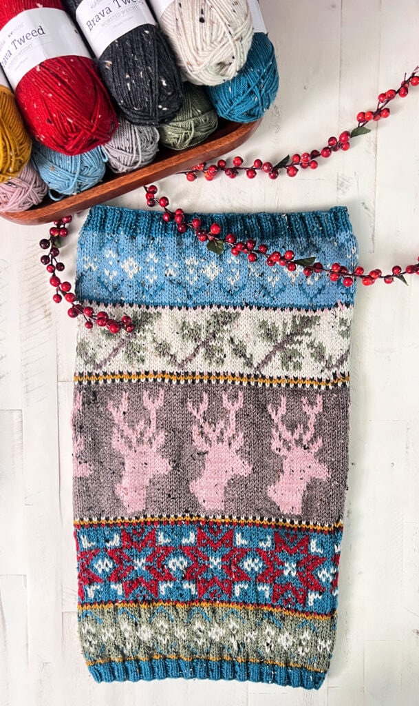 A colorful Dancer's Delight Knit Cowl with patterns of reindeer, snowflakes, and trees lies on a white surface, next to skeins of yarn in various colors and a string of red berries, ready for its final edging. -Marly Bird