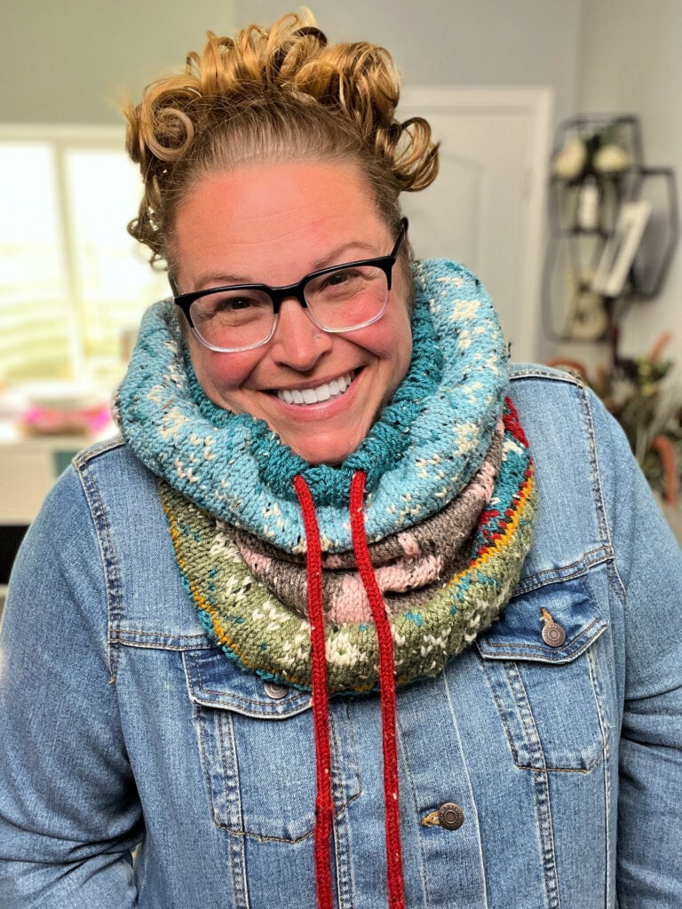 A smiling person with curly hair and glasses wears a blue denim jacket and a colorful, chunky dancer's delight knit cowl with red drawstrings, featuring a bold final edging, standing indoors in a well-lit room. -Marly Bird