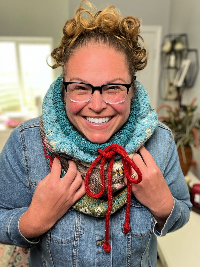 A smiling person with curly hair and glasses wears a colorful Dancer's Delight Knit Cowl with red ties and a stylish final edging over a denim jacket, standing indoors with bright windows and plants in the background. -Marly Bird