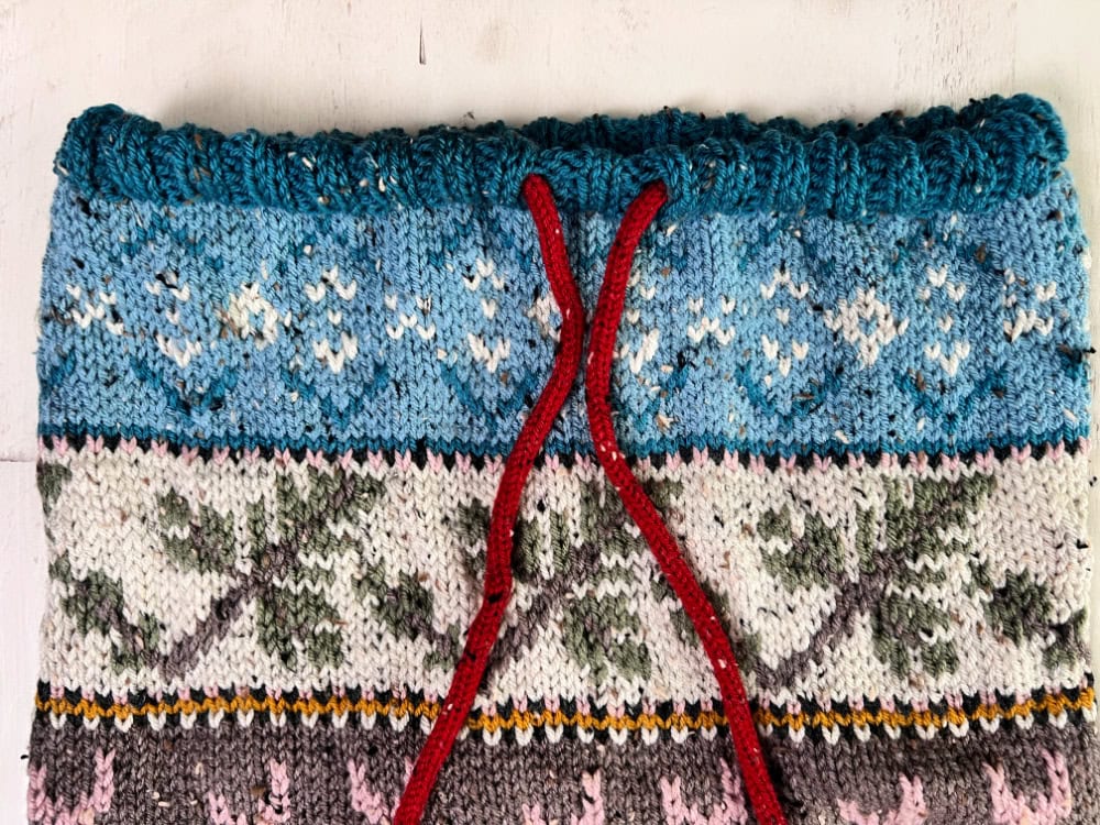 A close-up of the Dancer's Delight Knit Cowl showcases blue, white, green, and gray patterns with geometric and floral motifs, finished with a red drawstring threaded through the final edging. -Marly Bird