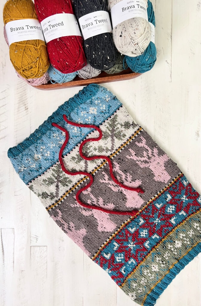 A colorful knitted piece featuring intricate patterns and reindeer designs lies on a white surface, surrounded by skeins of Brava Tweed yarn. Two red yarn strings hint at the final edging for this Dancer's Delight Knit Cowl. -Marly Bird