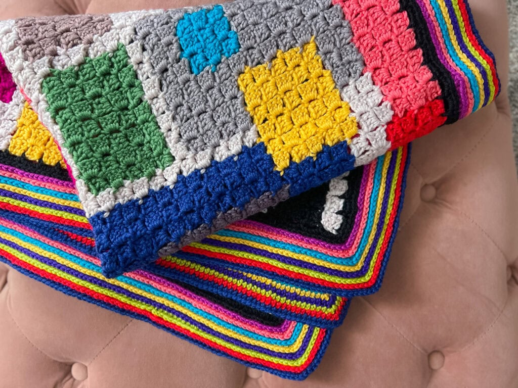 A FREE Crochet Chic Sheep Blanket with colorful blocks and stripes in blue, green, yellow, red, pink, and gray is folded neatly on a soft pink, tufted cushion. -Marly Bird