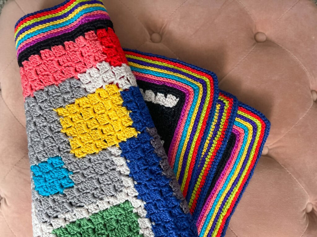 A colorful FREE Crochet Chic Sheep Blanket with various textured squares and a multicolored striped border, draped over a tufted, light pink cushion. -Marly Bird