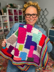 Person with glasses displays a geometric-pattern crocheted blanket, showing stitch texture in a yarn-filled craft room.