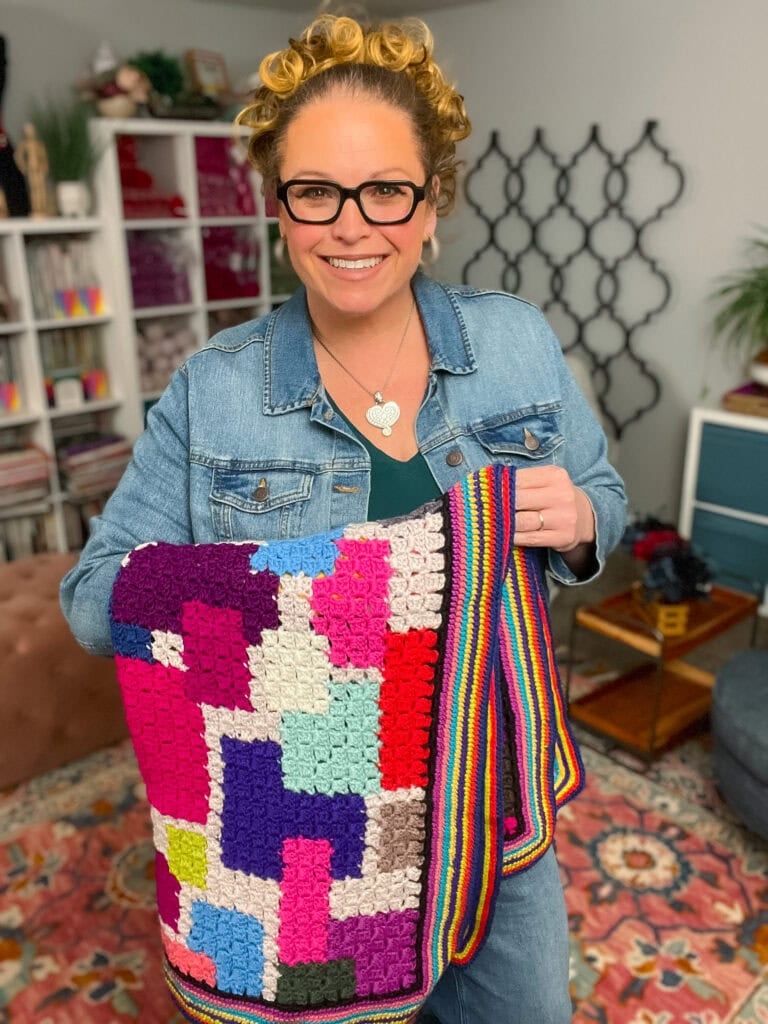 Woman displays a vibrant geometric crochet blanket in a cozy yarn-filled craft room, highlighting stitch detail and texture.