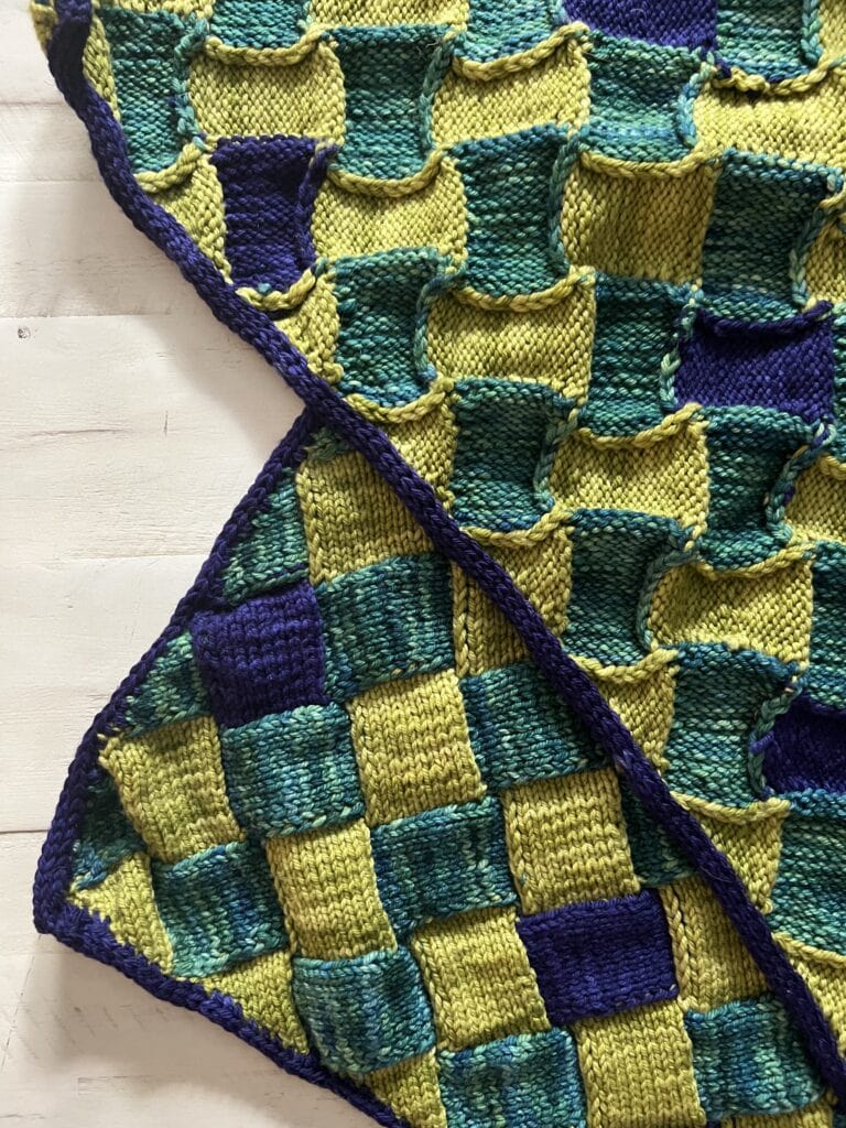 A close-up of a knitted blanket with a checkerboard pattern in green, yellow, and blue squares. The blanket is partially folded, showing the textured, raised design and neat edging. The background is a light wooden surface. -Marly Bird