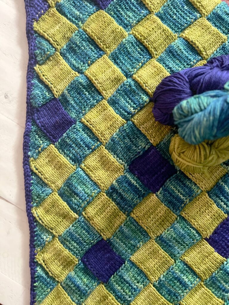 Crocheted checkerboard blanket in blue, green, and yellow with three matching skeins of yarn on a white wood background.