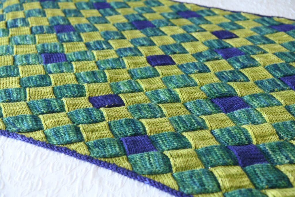 A close-up of a handwoven fabric featuring a checkerboard pattern in shades of green, yellow, and blue with a purple border, displayed on a textured white surface. -Marly Bird