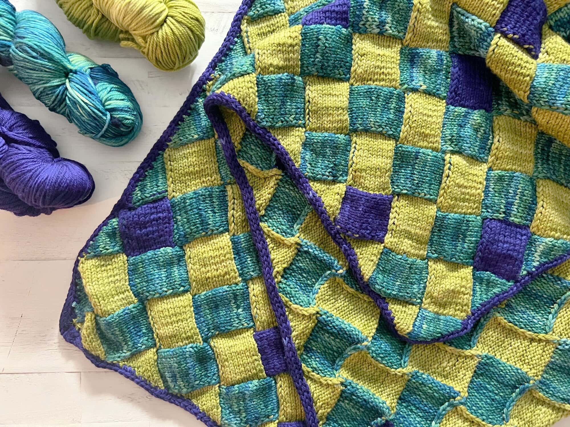 Hand-knitted checkered blanket in blue, green, and purple with visible texture, beside three matching yarn skeins on white.