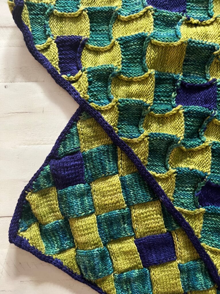 A close-up of a colorful knitted blanket with a geometric checkerboard pattern in yellow, green, and blue squares—edged with a purple border—displayed on a light wooden surface, perfect inspiration for free knitting and crochet patterns. -Marly Bird
