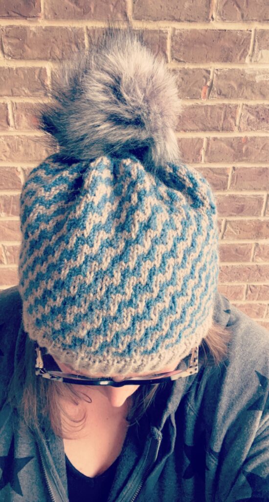 Top view of the cozy mosaic knit beanie on on Marly Bird. Image is against a brick wall background. Dynamic Wave Mosaic Knit Hat Pattern by Marly Bird in blue and light gray color and gray pompom - Dynamic Wave Mosaic Knit Hat Pattern by Marly Bird