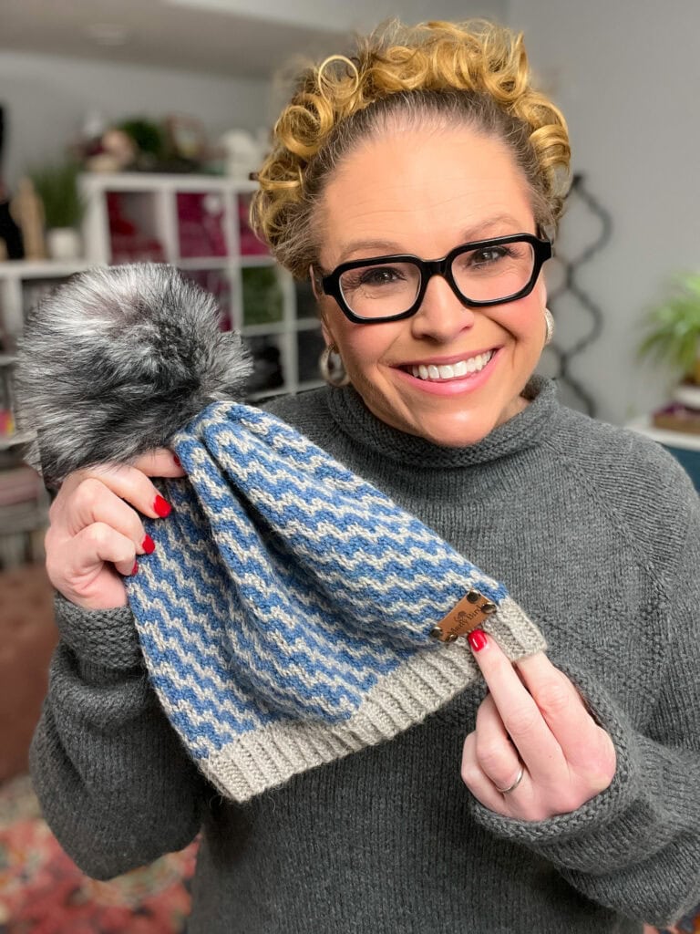 A smiling woman with curly hair and glasses holds up a blue and gray Mosaic Knit Hat Pattern with a pom-pom. She wears a gray sweater and red nail polish. Shelves and plants are visible in the background. -Marly Bird