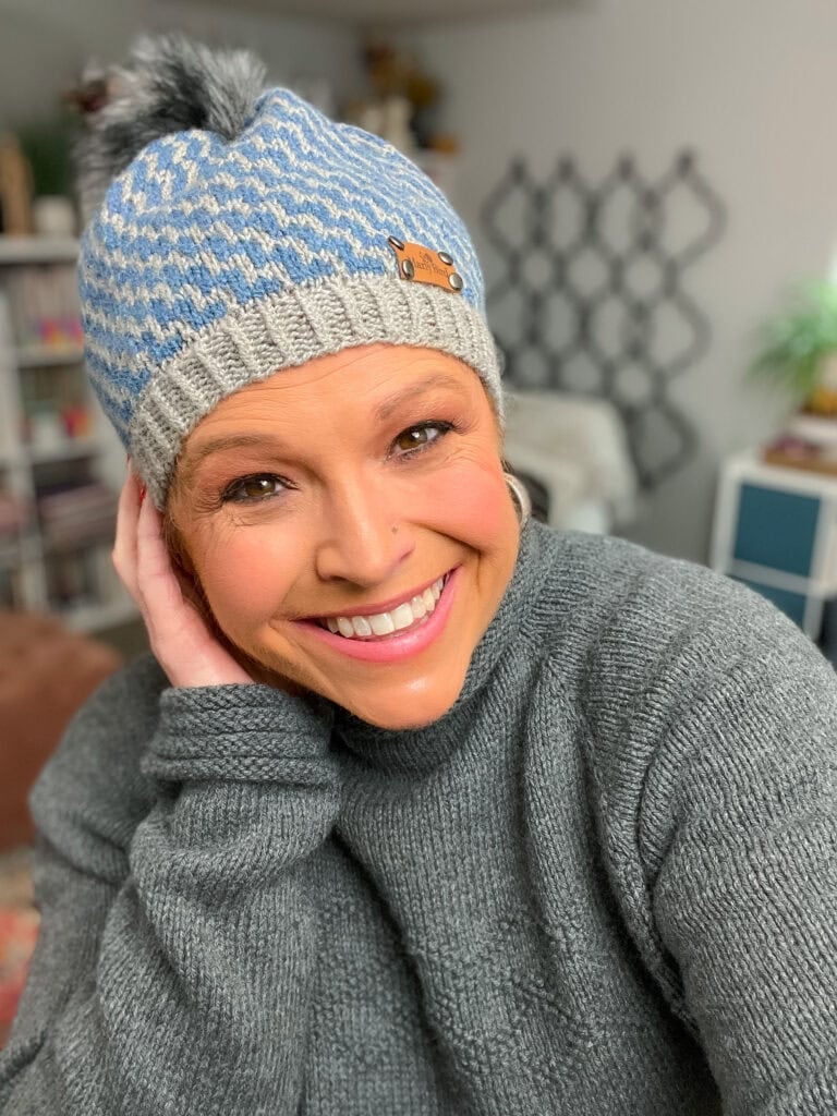 Blue and gray knit beanie with pom-pom, shown worn indoors, highlights textured stitches and colorwork; gray sweater also visible.