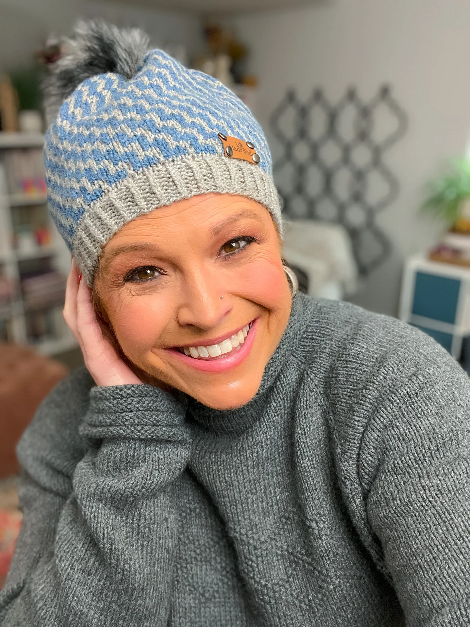 Blue and gray knit beanie with pom-pom, shown worn indoors, highlights textured stitches and colorwork; gray sweater also visible.