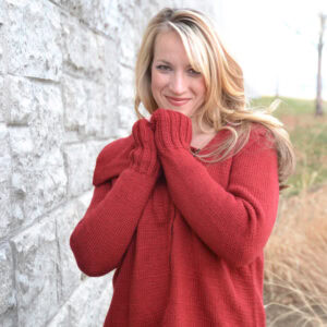 Woman wears a red Tunisian crochet sweater and matching mittens, showing intricate stitch detail outdoors by a stone wall.