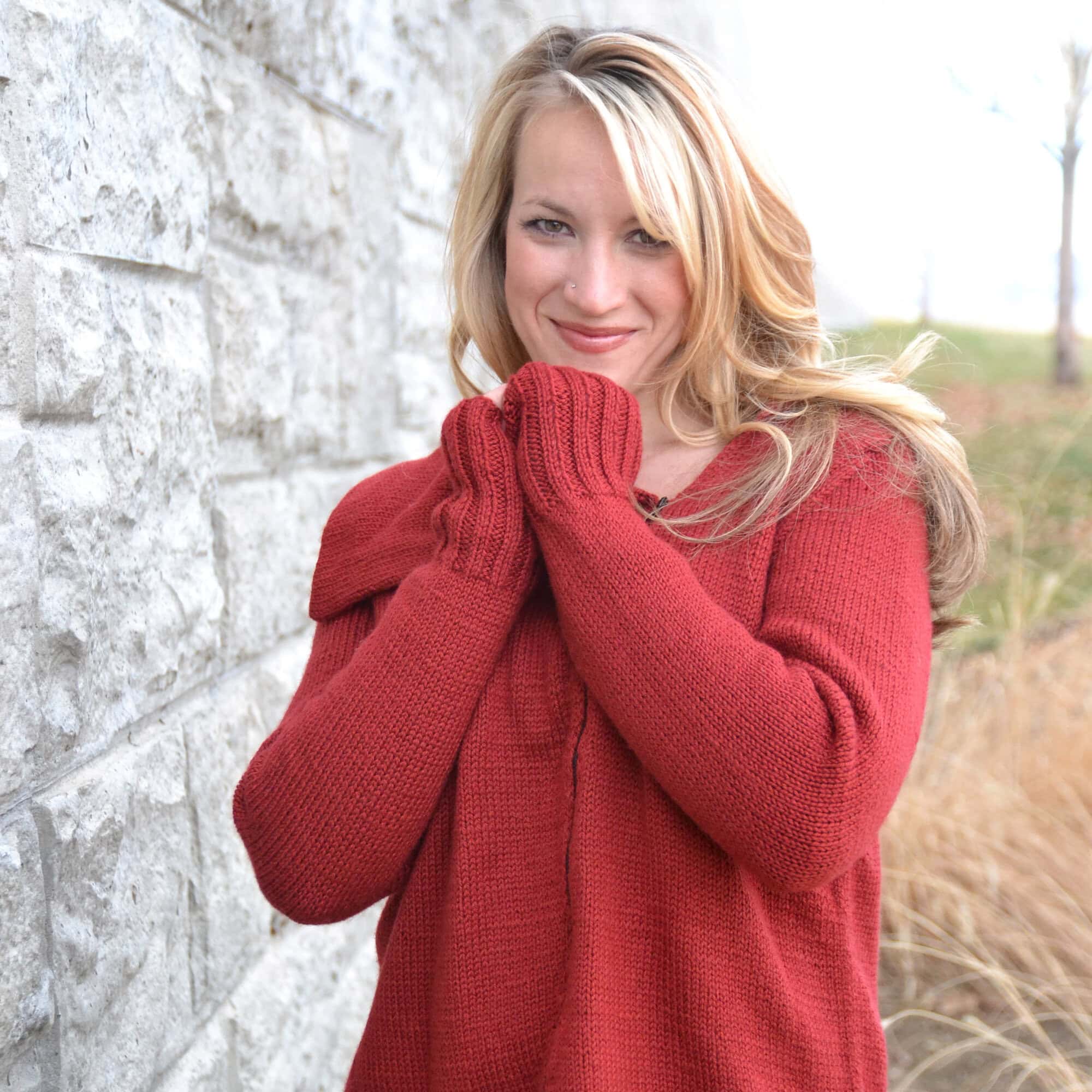 Woman wears a red Tunisian crochet sweater and matching mittens, showing intricate stitch detail outdoors by a stone wall.